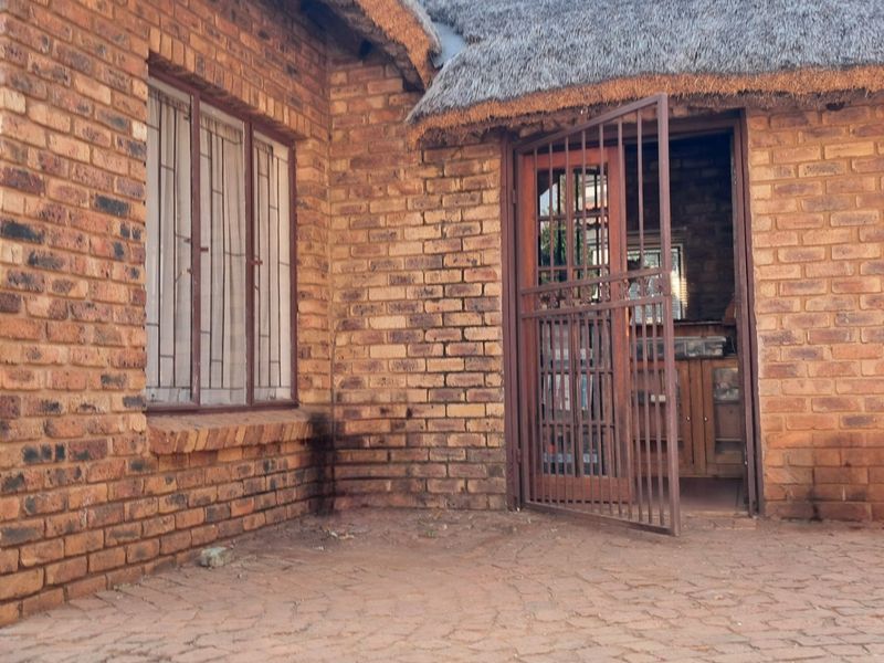 A rear exterior view of a face brick house featuring a traditional thatch roof, paved patio area, and secure metal entrance gate.