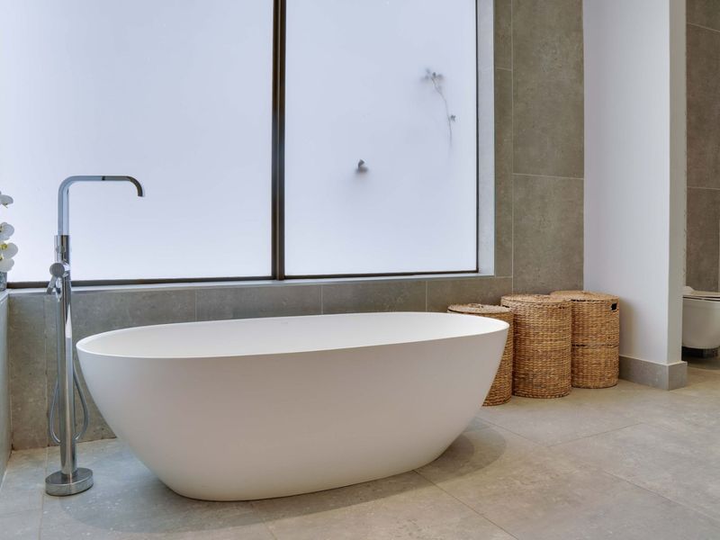 Modern bathroom featuring a sleek freestanding bathtub with floor-mounted mixer, large format tiling, and a wall-hung toilet under a large frosted window.