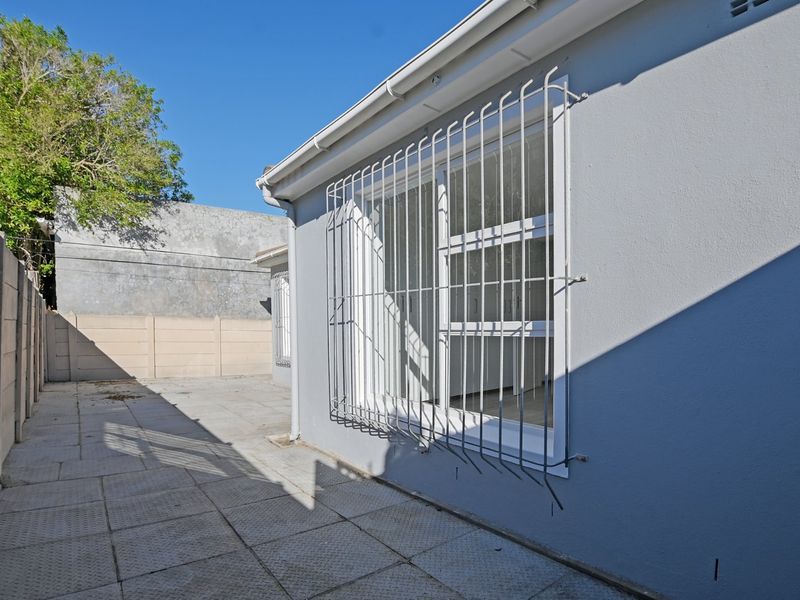 A view of the rear exterior area featuring a paved courtyard, boundary wall, and windows fitted with security bars.