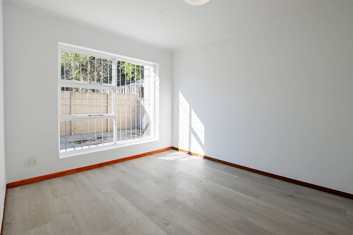 A bright, empty bedroom featuring light-toned laminate flooring, white walls, and a large window fitted with security bars.