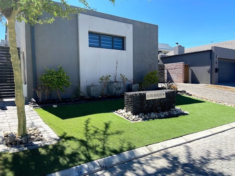 Modern front exterior featuring a well-maintained artificial lawn, stone-filled gabion feature wall, and paved driveway access.