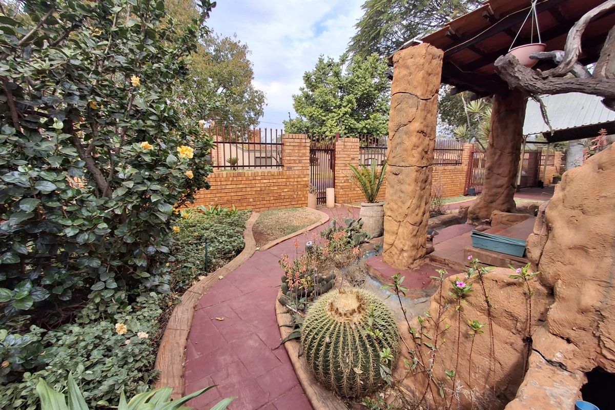 A rear garden area featuring a paved walkway, established landscaping, and a covered patio structure with textured concrete pillars and brick boundary walls.