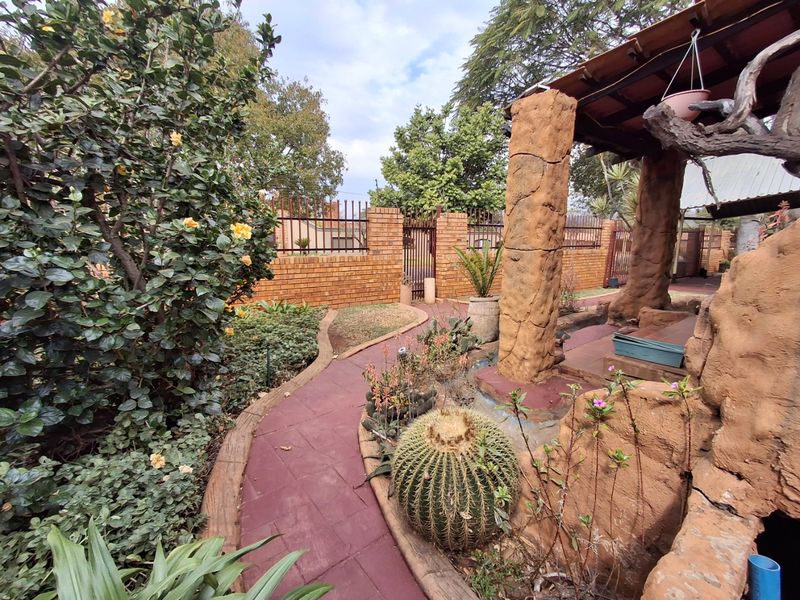 A rear garden area featuring a paved walkway, established landscaping, and a covered patio structure with textured concrete pillars and brick boundary walls.