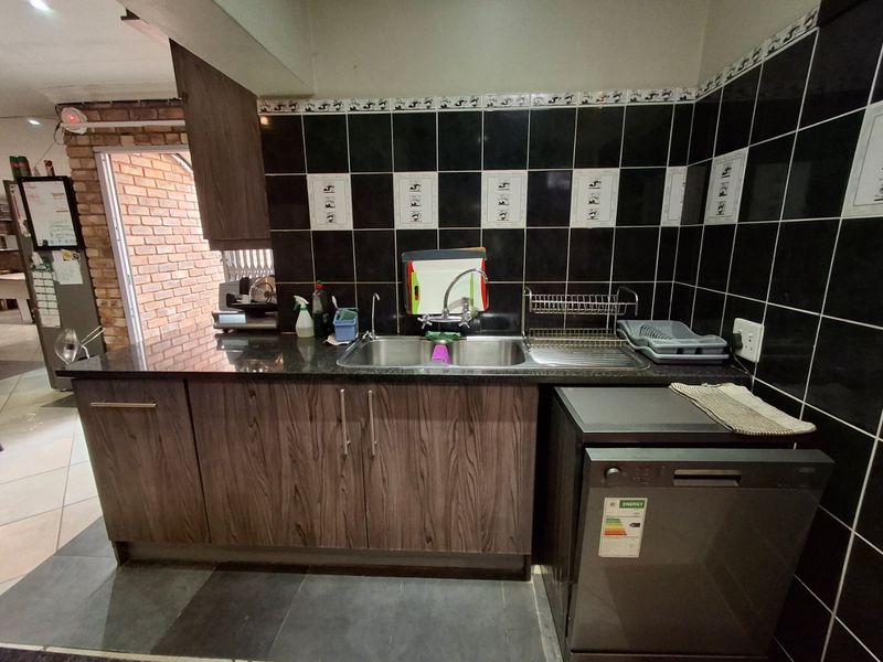 A functional scullery area featuring dark granite countertops, wood-grain cabinetry, black tiled splashbacks, and a double bowl stainless steel sink.