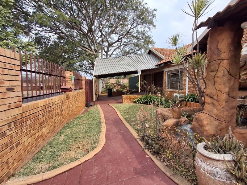 Front exterior view featuring a paved walkway, covered carport, brick boundary wall with security fencing, and a decorative rock water feature.