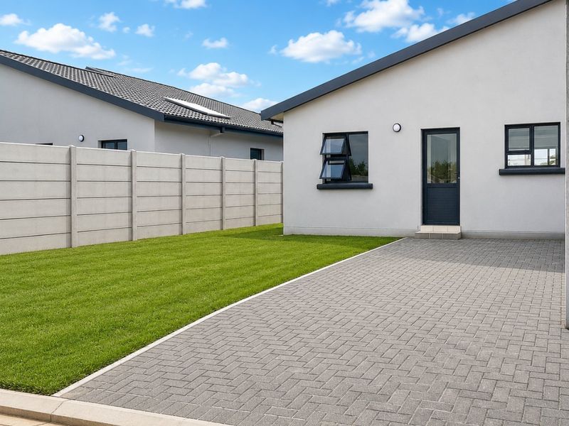 A well-maintained rear exterior view featuring a paved driveway, a neat lawn, and a secure precast concrete boundary wall.
