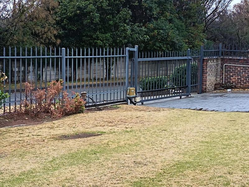 Front entrance view featuring a metal security gate with an electric motor, brick boundary wall, and paved driveway access.