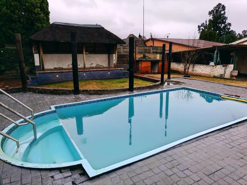 Outdoor pool area featuring a swimming pool with integrated steps and a thatched lapa structure, set within a paved backyard space.