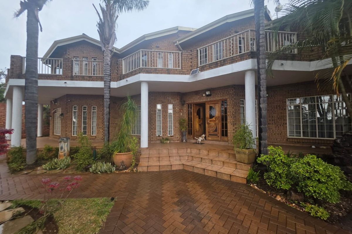 A two-story brick house featuring a spacious tiled patio, upper-level balcony, and a paved driveway entrance.