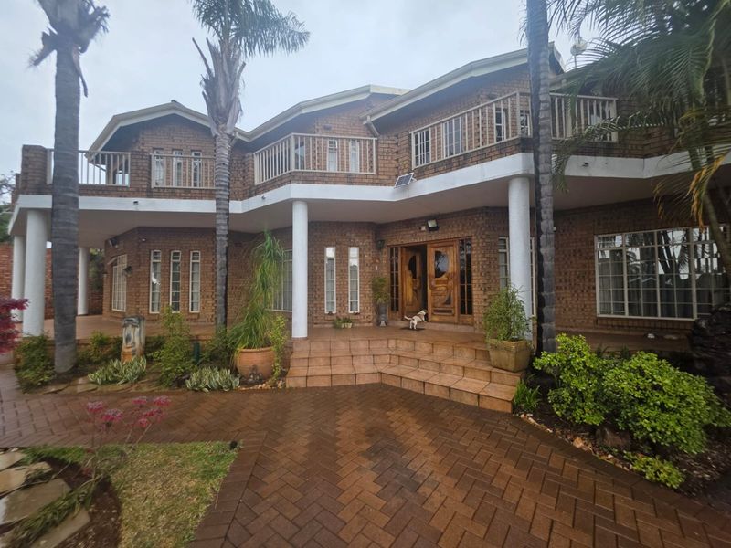 A two-story brick house featuring a spacious tiled patio, upper-level balcony, and a paved driveway entrance.