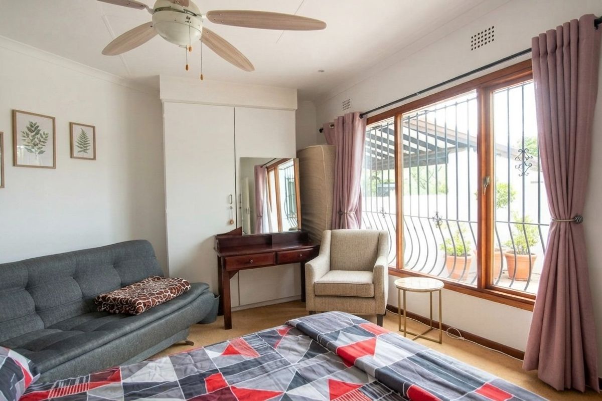 A bedroom featuring built-in wardrobes, a ceiling fan, and large windows with security bars, offering natural light and views of the outdoor area.