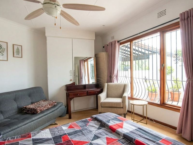 A bedroom featuring built-in wardrobes, a ceiling fan, and large windows with security bars, offering natural light and views of the outdoor area.