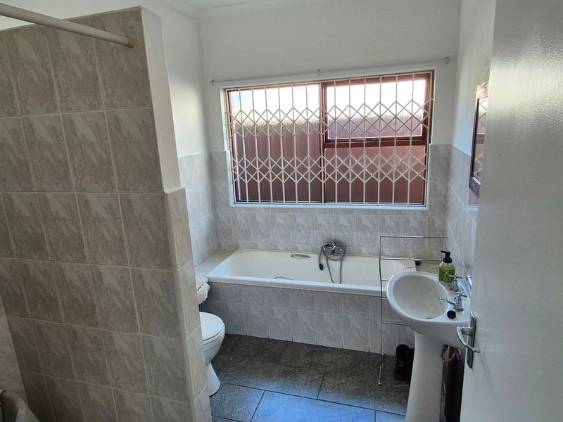 A functional bathroom featuring a tiled shower enclosure, a built-in bathtub, a pedestal washbasin, and a toilet, with security bars installed on the window.