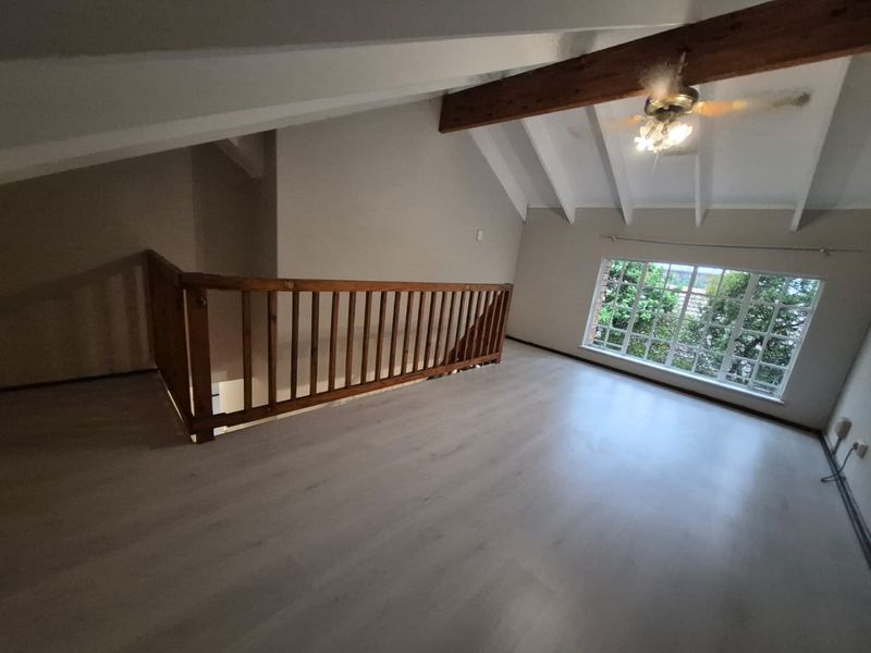 Spacious loft-style living area featuring vaulted ceilings with exposed timber beams, light laminate flooring, and a large window providing natural light.