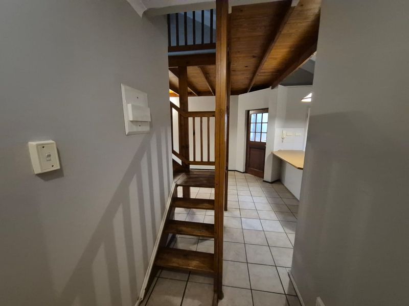 A hallway featuring a wooden staircase with balustrade, exposed timber ceiling beams, and tiled flooring, leading towards a kitchen area and exterior door.