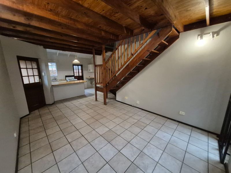 Open-plan living area featuring exposed timber ceiling beams, a wooden staircase, and tiled flooring, leading into a functional kitchen space.