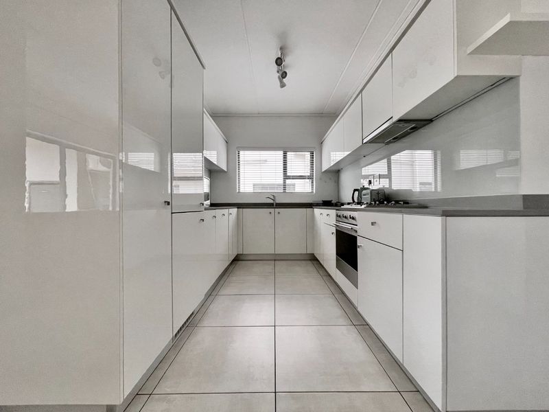 A modern, galley-style kitchen featuring sleek white cabinetry, stone countertops, integrated appliances, and large format floor tiles.