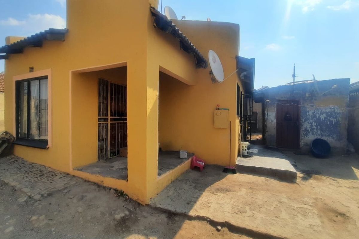 Rear view of a yellow-painted house featuring a small covered patio area, security gates, and an adjacent outbuilding with weathered exterior walls.