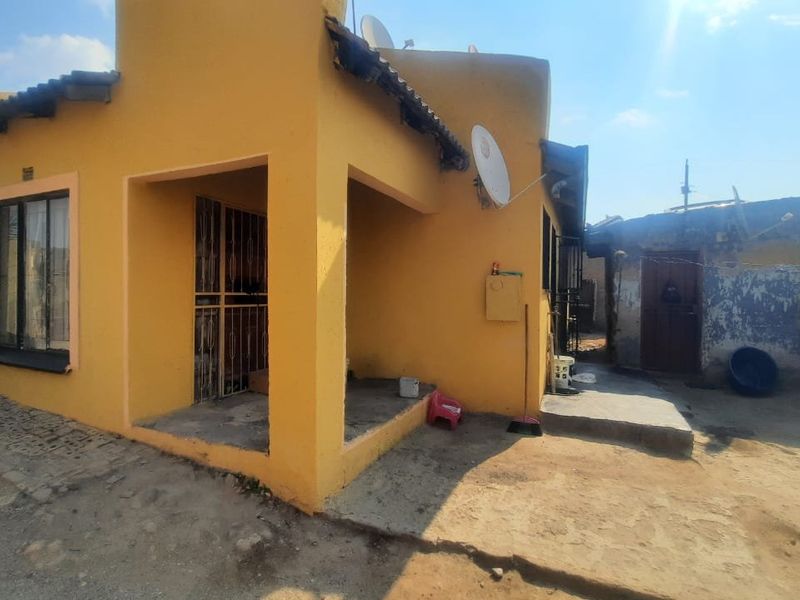 Rear view of a yellow-painted house featuring a small covered patio area, security gates, and an adjacent outbuilding with weathered exterior walls.