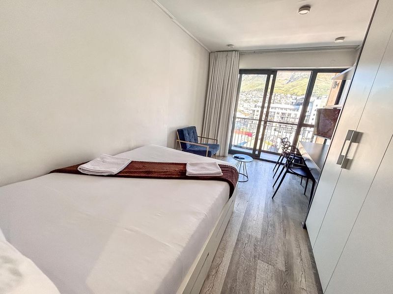 A bright bedroom featuring laminate flooring, a built-in wardrobe, and a sliding glass door leading to a balcony with city and mountain views.
