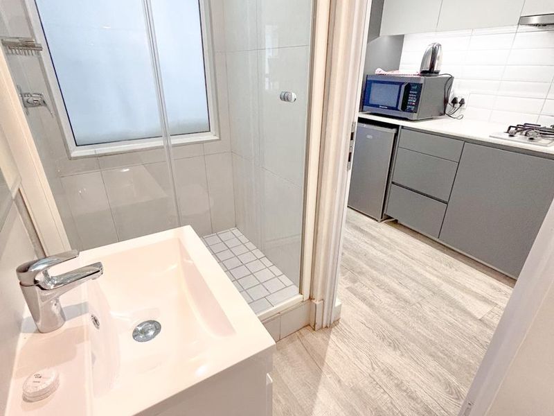 Modern bathroom featuring a vanity basin, glass-enclosed shower, and toilet, with adjacent view into a compact kitchen area.