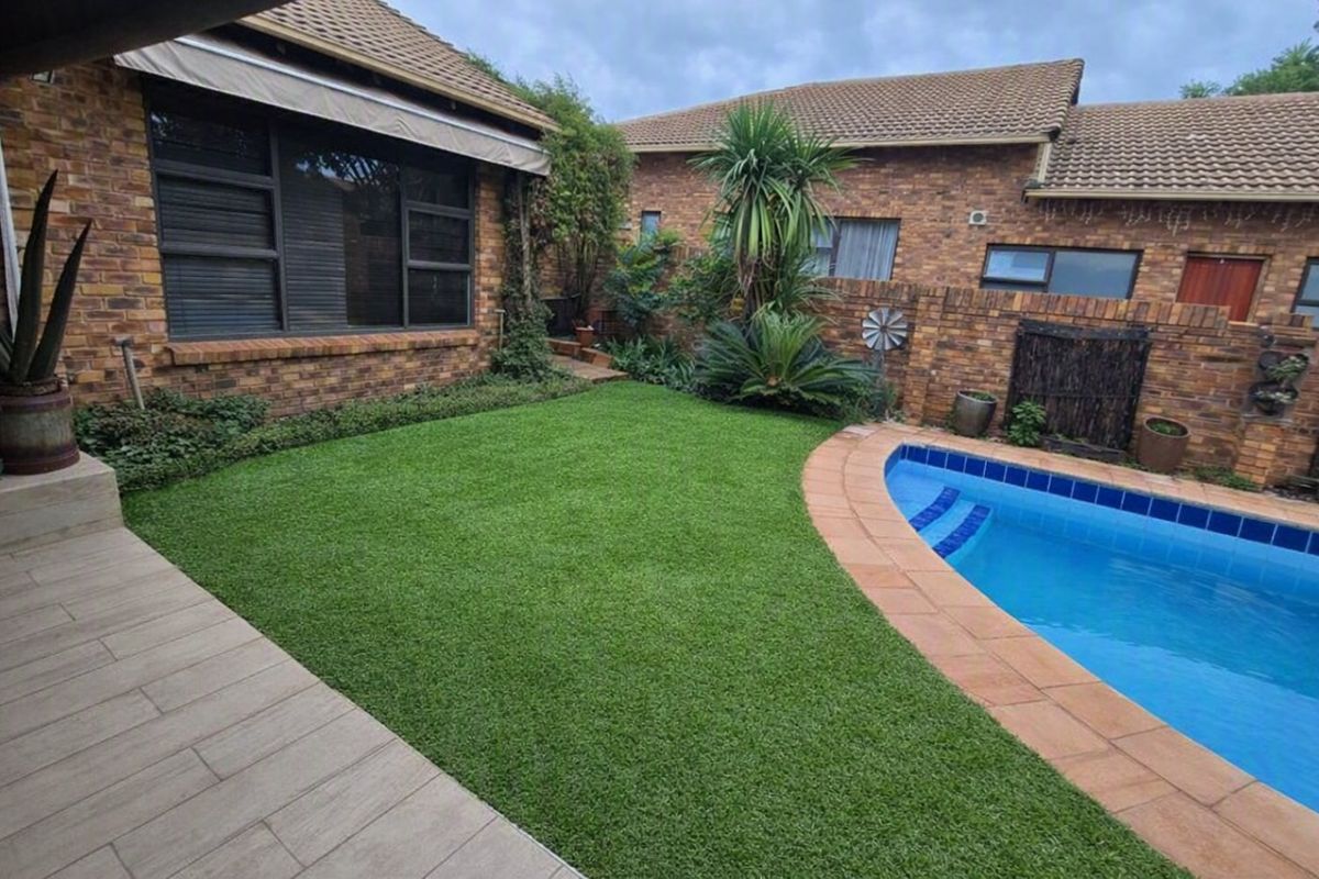 A well-maintained outdoor pool area featuring a swimming pool, artificial lawn, tiled patio, and face-brick exterior walls.