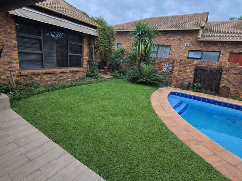 A well-maintained outdoor pool area featuring a swimming pool, artificial lawn, tiled patio, and face-brick exterior walls.