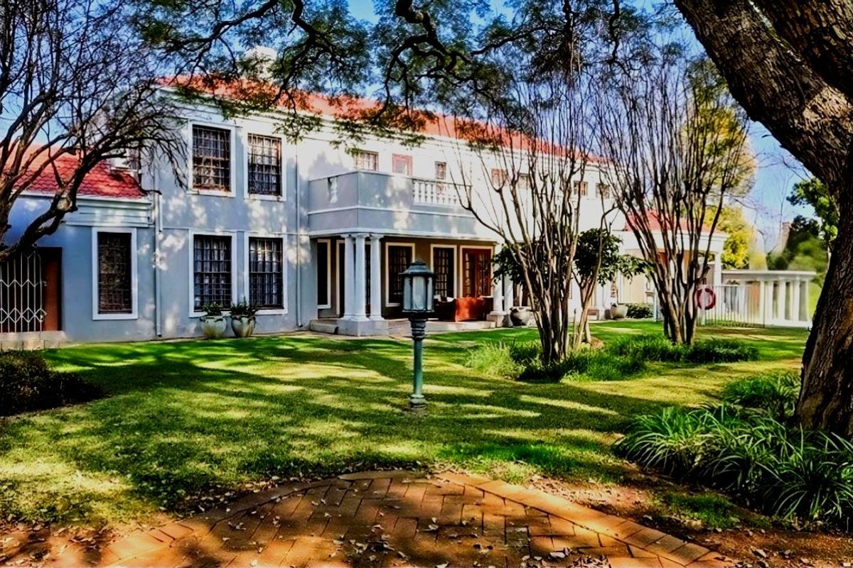 A spacious rear garden view of a two-story residence featuring a terracotta tiled roof, a balcony with columns, and a brick-paved patio area.