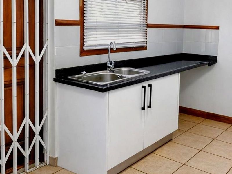 A clean and functional scullery area featuring a double sink, dark laminate countertops, white base cabinetry, and neutral tiled flooring.