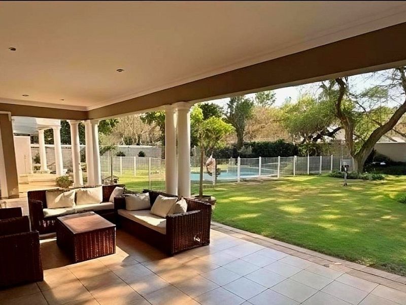 Spacious covered patio featuring tiled flooring and structural columns, overlooking a well-maintained garden and swimming pool area.