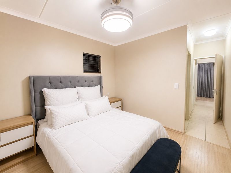 A well-maintained bedroom featuring laminate flooring, neutral wall tones, and a modern ceiling light fixture, with an adjoining hallway visible.