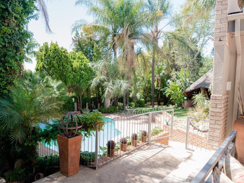 A lush backyard featuring a swimming pool, a brick-paved patio area, and a traditional thatched lapa surrounded by mature trees and garden landscaping.