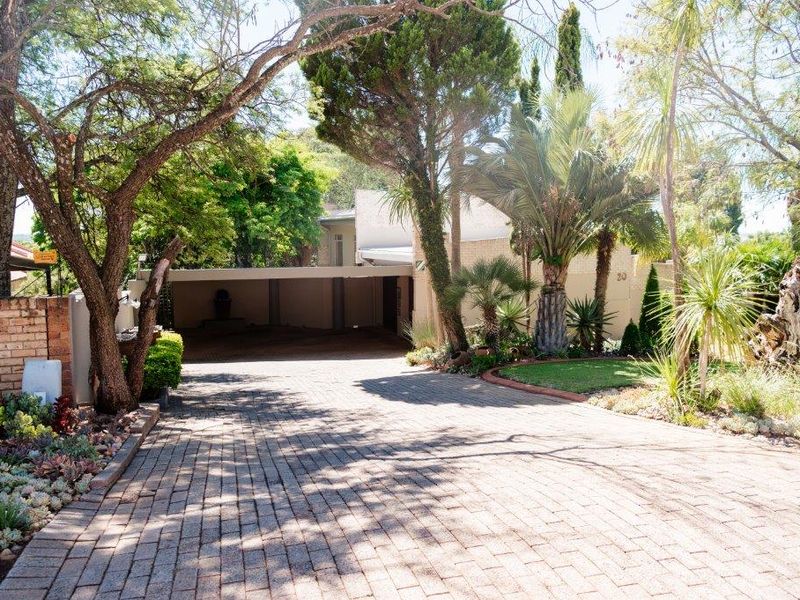 Front view of the property featuring a brick paved driveway leading to a carport, surrounded by a mature landscaped garden.