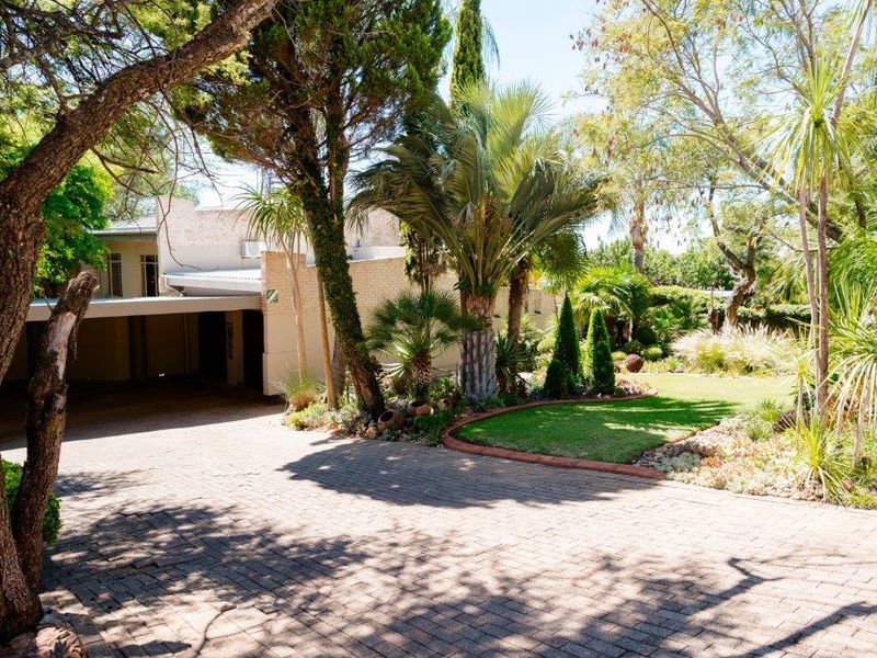 A well-maintained front exterior featuring a paved driveway, covered carport, and established landscaped garden with mature trees and shrubs.