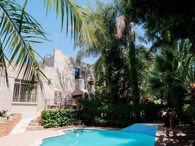 A private outdoor pool area featuring a paved patio, built-in fireplace, and mature garden surroundings, set against the rear brick facade of the house.