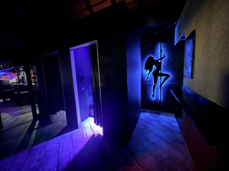 Interior view of a commercial bar area featuring tiled flooring, dark wall partitions, and decorative neon-style accent lighting.