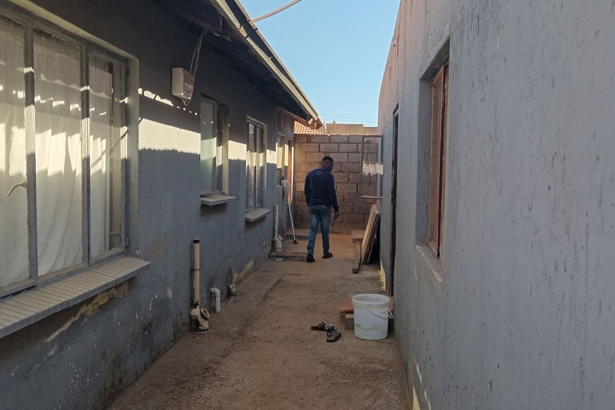 A narrow exterior side passage between two building structures featuring concrete pathways and boundary walls requiring maintenance and surface repairs.