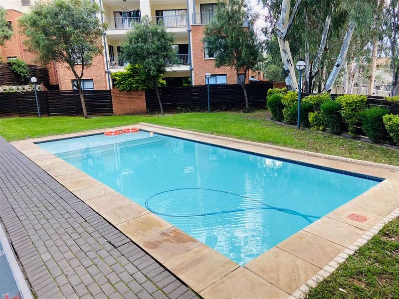 A well-maintained communal swimming pool area featuring paved surrounds, established garden landscaping, and perimeter fencing within an apartment complex setting.