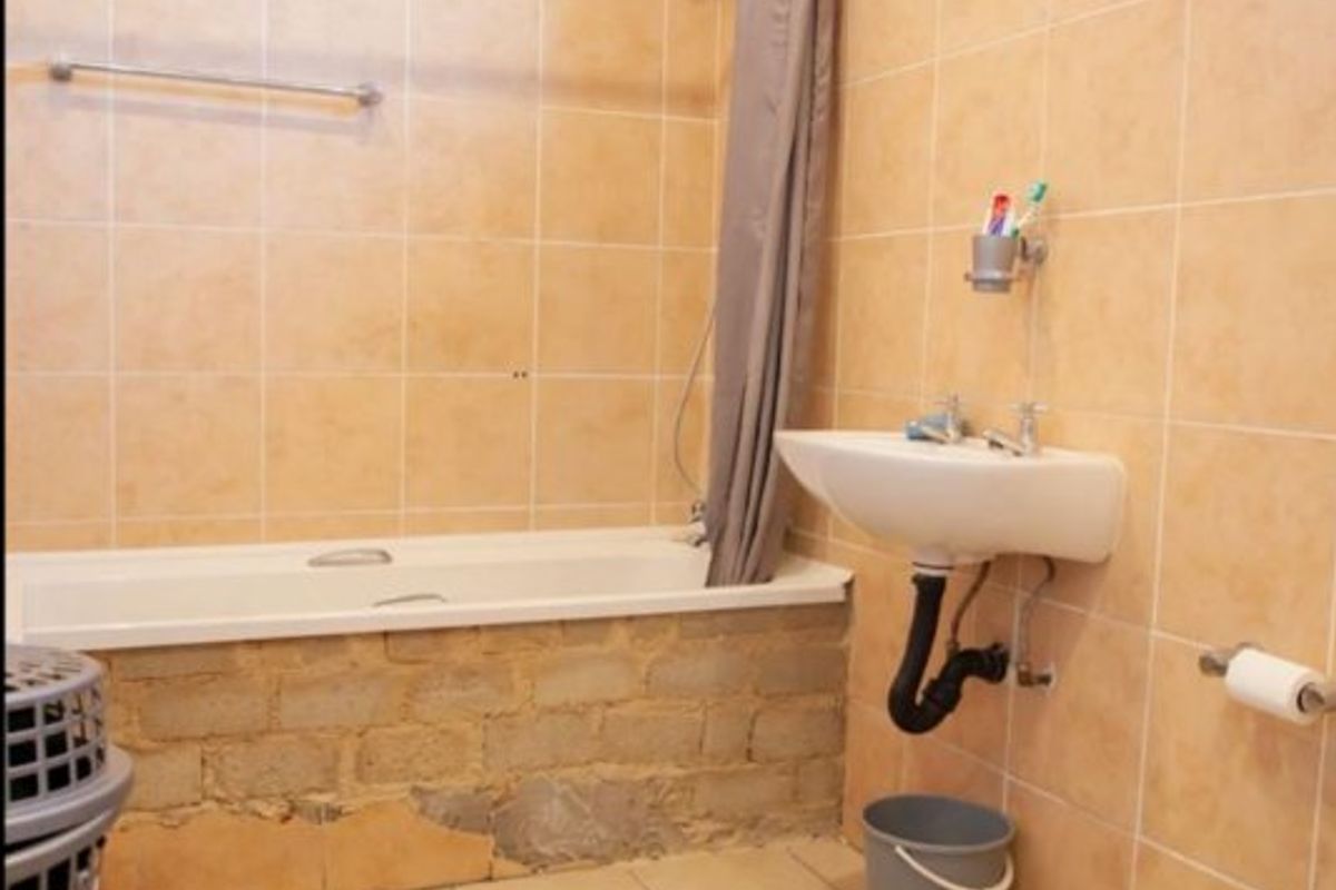 A bathroom featuring a bathtub, wall-mounted basin, and toilet, with tiled finishes requiring repair and cosmetic updates to the bathtub apron and plumbing.