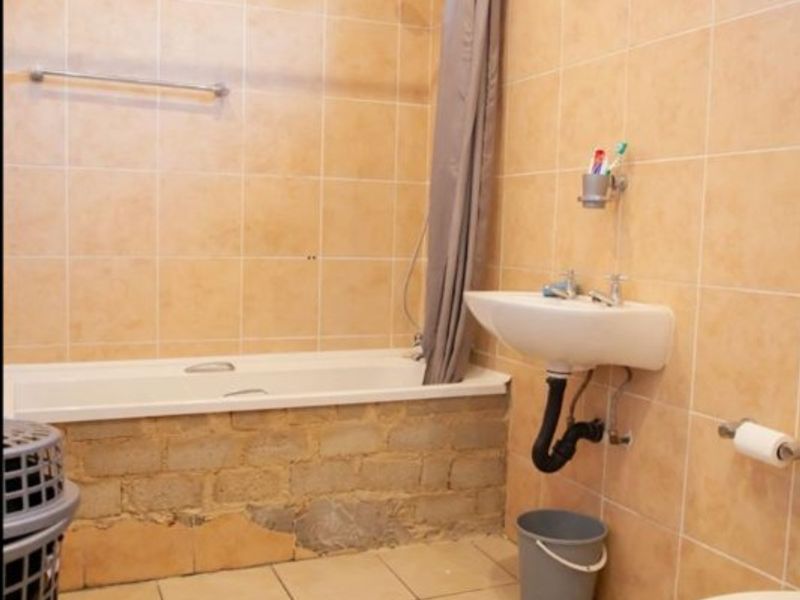 A bathroom featuring a bathtub, wall-mounted basin, and toilet, with tiled finishes requiring repair and cosmetic updates to the bathtub apron and plumbing.