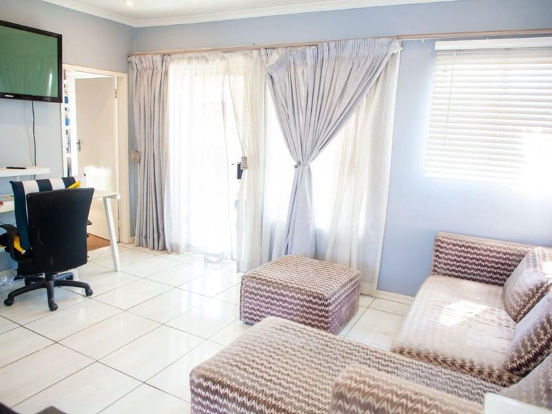 A bright living area featuring white tiled flooring, large sliding glass doors with curtains, and venetian blinds, offering a functional and well-lit space.