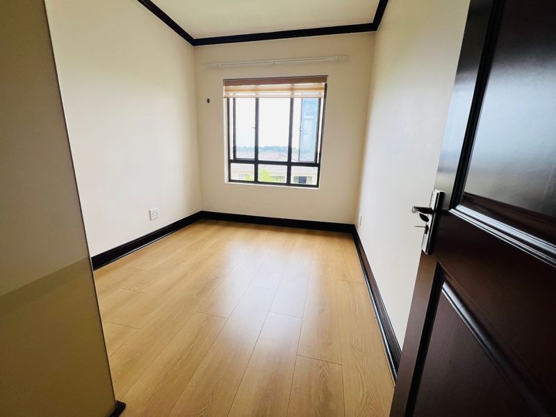 A bright, empty bedroom featuring light wood-look laminate flooring, dark contrasting skirting boards, and a window with a fitted roller blind.