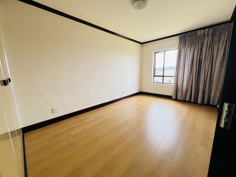 A bright, empty bedroom featuring light wood-look laminate flooring, dark trim accents, and a window with curtains.