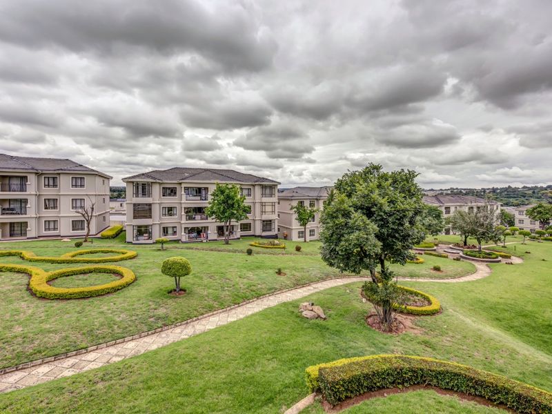A wide-angle view of the apartment complex grounds, featuring well-maintained landscaped gardens, paved pathways, and multi-story residential buildings under a cloudy sky.