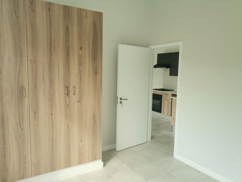 A bright, empty bedroom featuring modern wood-finish built-in wardrobes, neutral tiled flooring, and a view into the adjacent kitchen area.