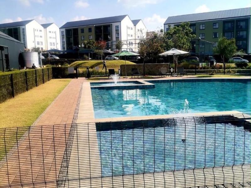 Communal swimming pool area featuring a water fountain, paved surround, and landscaped garden within a secure apartment complex.