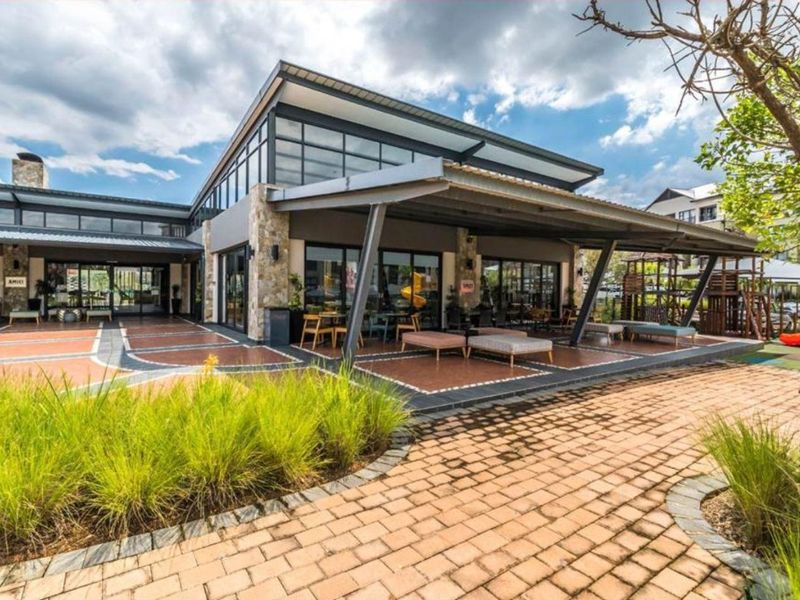 A modern clubhouse exterior featuring a spacious covered patio, stone accents, and large glass doors opening onto a paved terrace area.