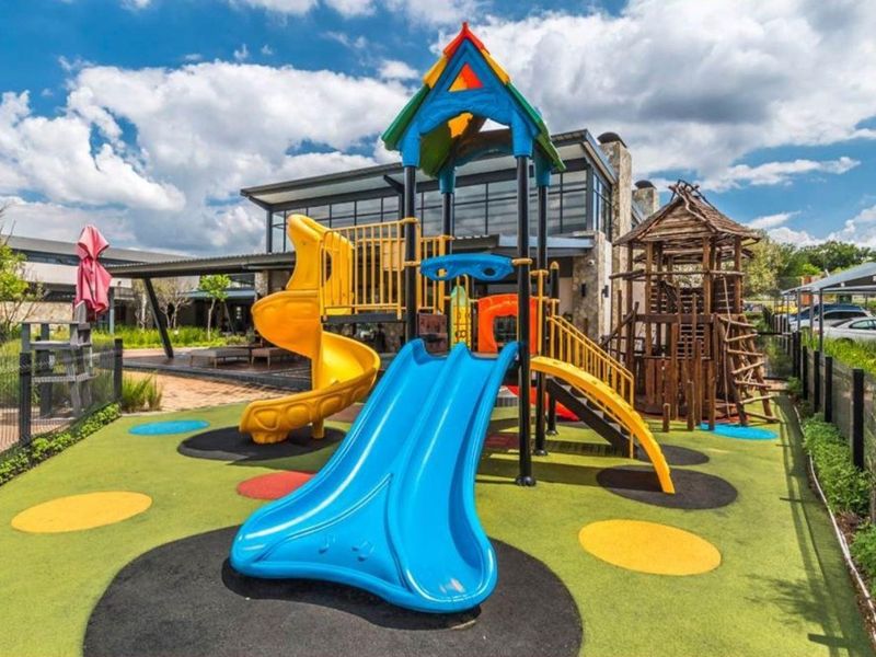 A well-maintained outdoor playground area featuring colorful play structures and safety flooring, situated in the rear communal space of the apartment complex.