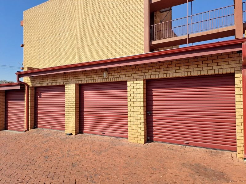 A row of secure, red roller shutter garage doors set against a yellow brick building with a paved driveway.