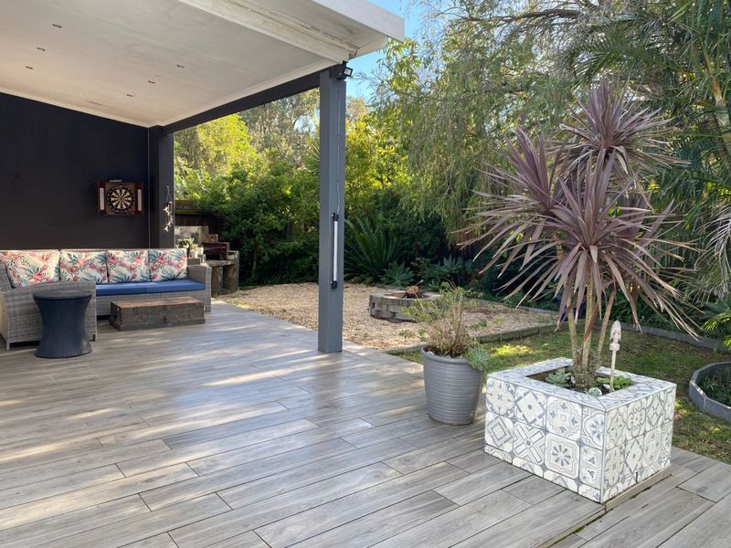 A spacious covered patio featuring wood-look tiled flooring, recessed lighting, and a built-in braai, overlooking a landscaped garden with a stone fire pit.